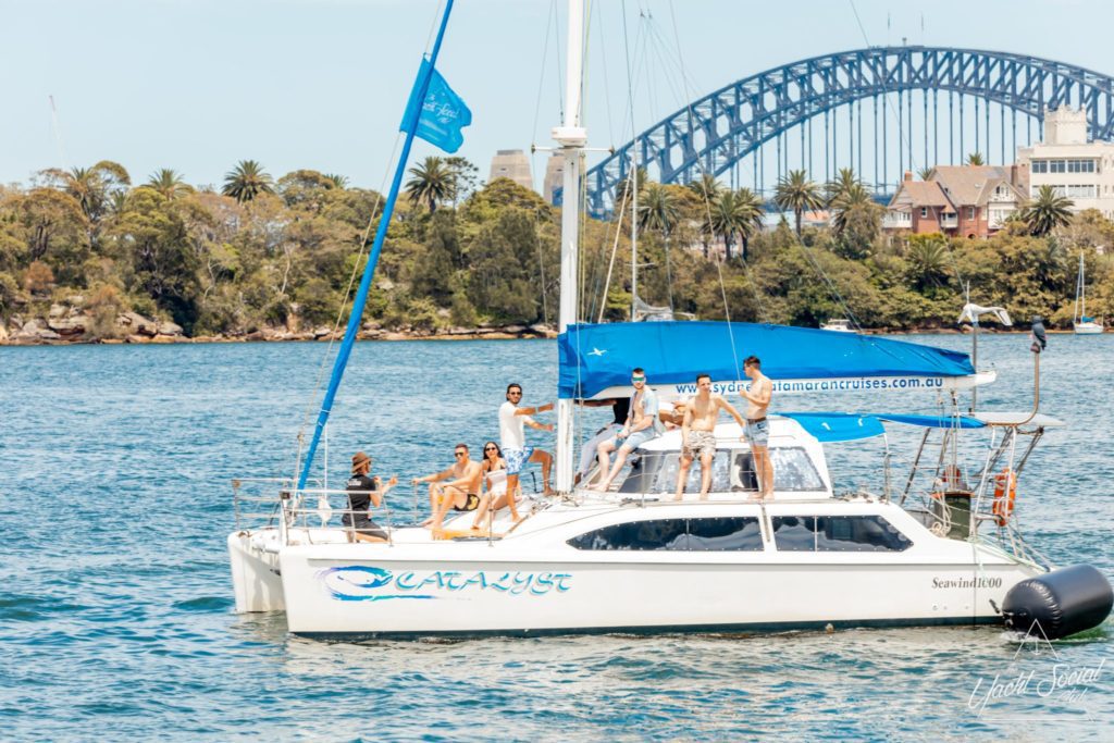 A group of people are having a great time on the Catalyst catamaran in a harbor where the Sydney Harbour Bridge is visible in the background, making it ideal for anyone seeking an unforgettable Catamaran party experience in Sydney.