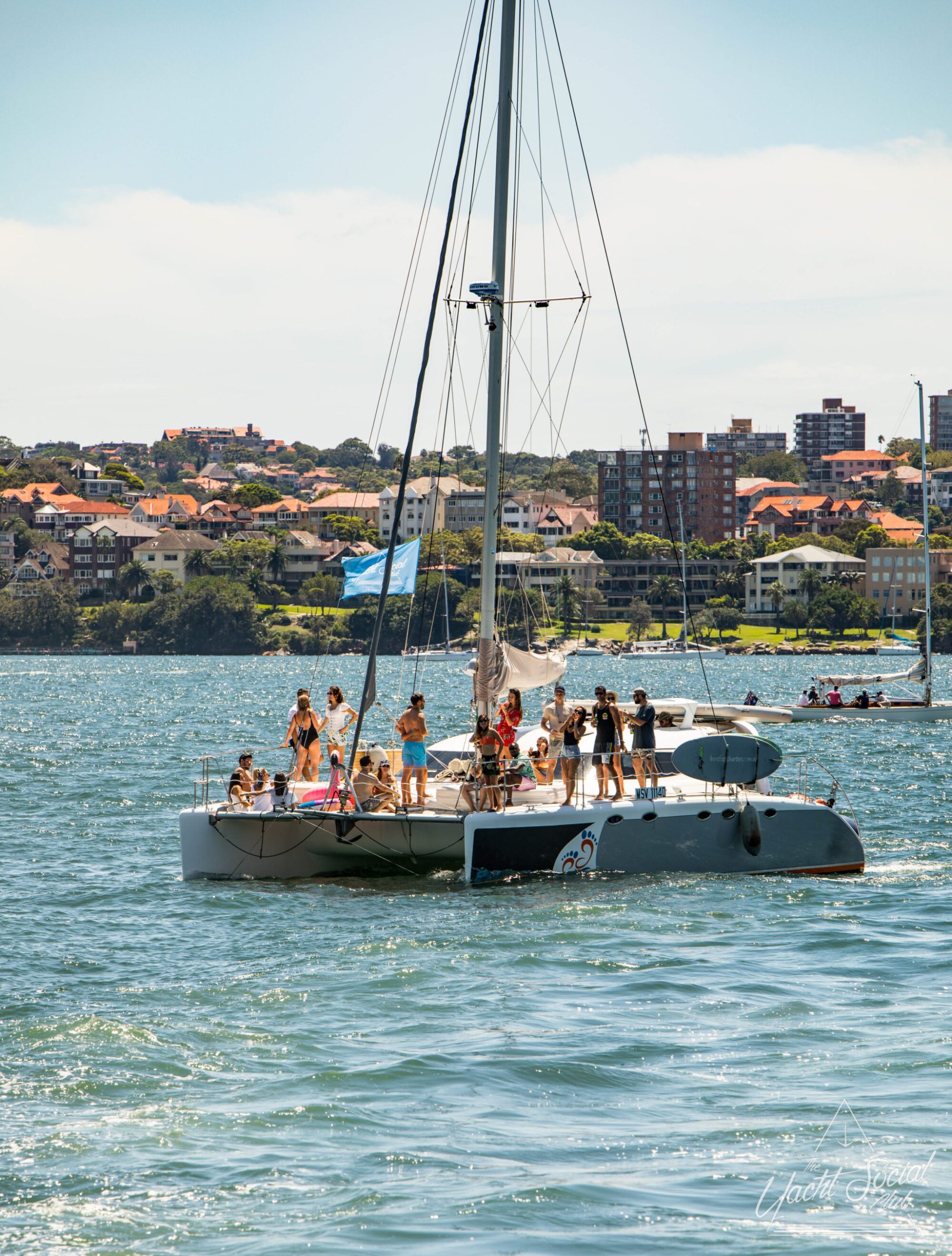 A group of people are gathered on a luxury yacht in a body of water, with a coastline featuring residential buildings in the background.