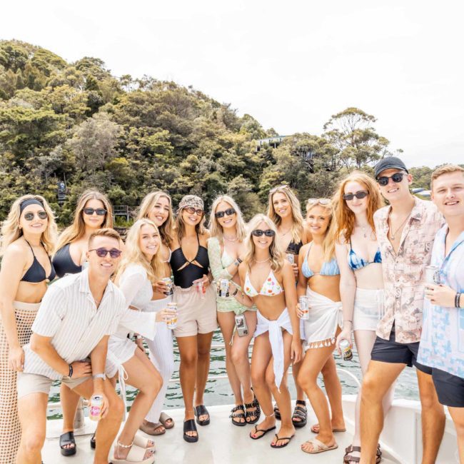 A group of people in casual summer attire and swimwear pose together on a boat by a body of water, with a wooded shore in the background, enjoying their private yacht charter Sydney Harbour.