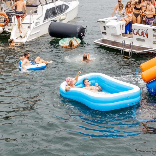 People are enjoying a lake party with inflatables and boats. Some float on inflatable beds while others are on boats or swimming. The scene includes a slide attached to a boat, perfect for those looking for a fun DJ boat hire in Sydney.
