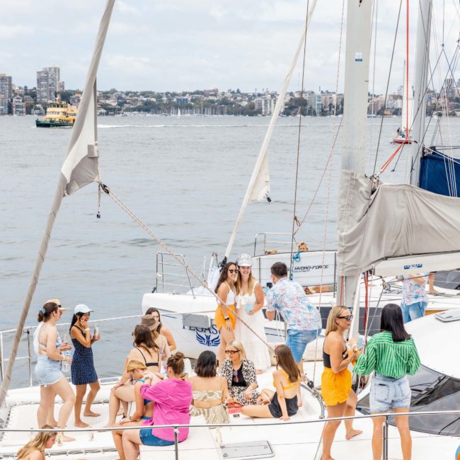A group of people socializing on a white sailboat in a city harbor setting, with buildings and another boat visible in the background. Perfect for Corporate boat events Sydney or Catamaran party Sydney, this scene offers an exquisite maritime escape against a picturesque urban backdrop.