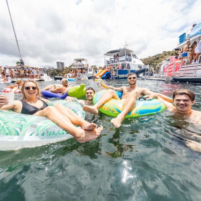 A group of people enjoy the water on inflatable floats in a busy marina area with boats and other individuals visible in the background, perfect for a Sydney boat party hire.