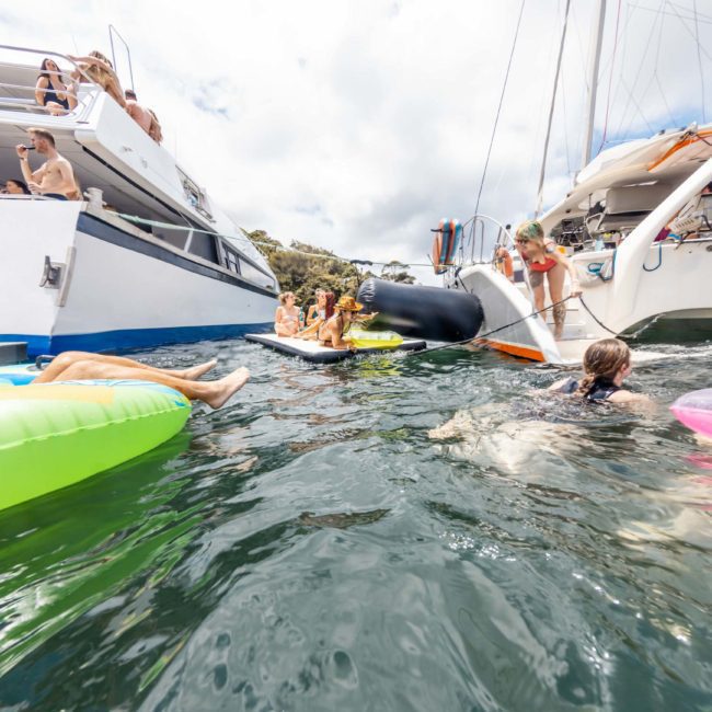 People enjoying a sunny day on and around boats and inflatables in the water, with some swimming and others relaxing on the decks of a luxury yacht hire Sydney.