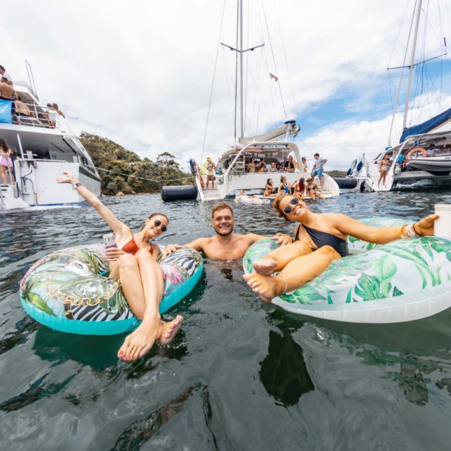 Three people float on inflatable rings in the water, smiling and raising their arms, with boats including a luxury yacht hire Sydney in the background.