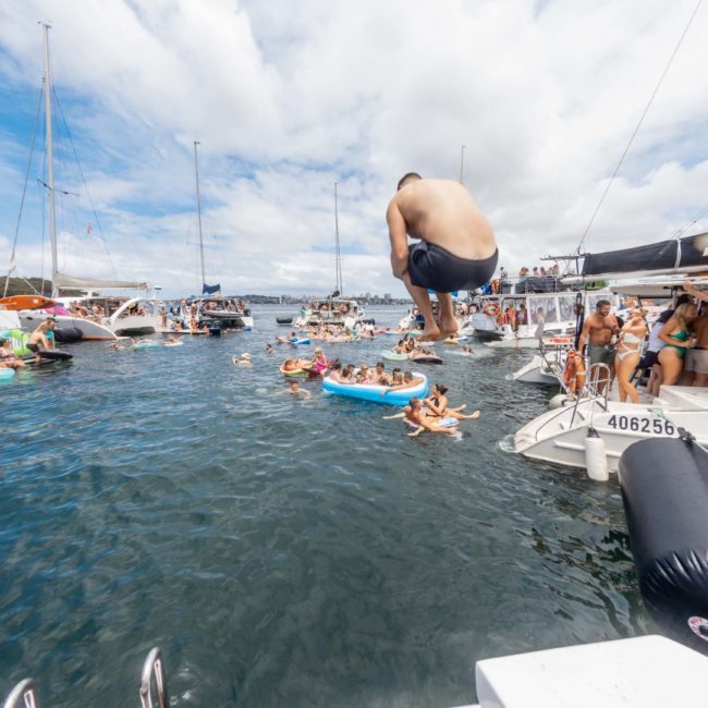 A man jumps into the water from a boat, surrounded by numerous boats with people swimming and enjoying the water on a sunny day. Nearby, a DJ boat hire in Sydney pumps out upbeat tunes, enhancing the festive atmosphere.
