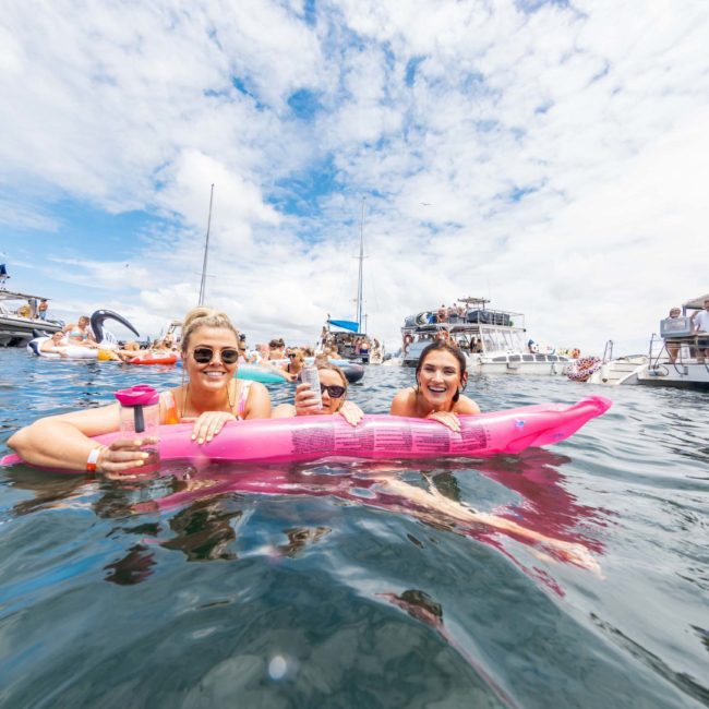 Three people are on a pink inflatable in a body of water, with boats and other people in the background under a partly cloudy sky. Experience the excitement of Sydney's waters, whether it's private yacht charters on Sydney Harbour or enjoying DJ boat hire events.