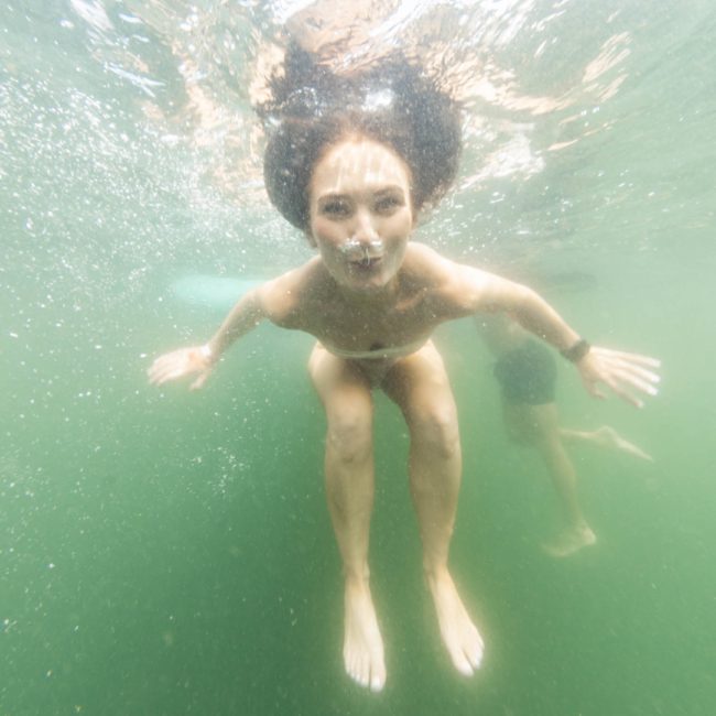 Underwater view of a woman swimming with another person partially visible in the background. Both are submerged in greenish water, and the woman is looking towards the camera, perhaps enjoying a break from a luxury yacht hire Sydney experience.