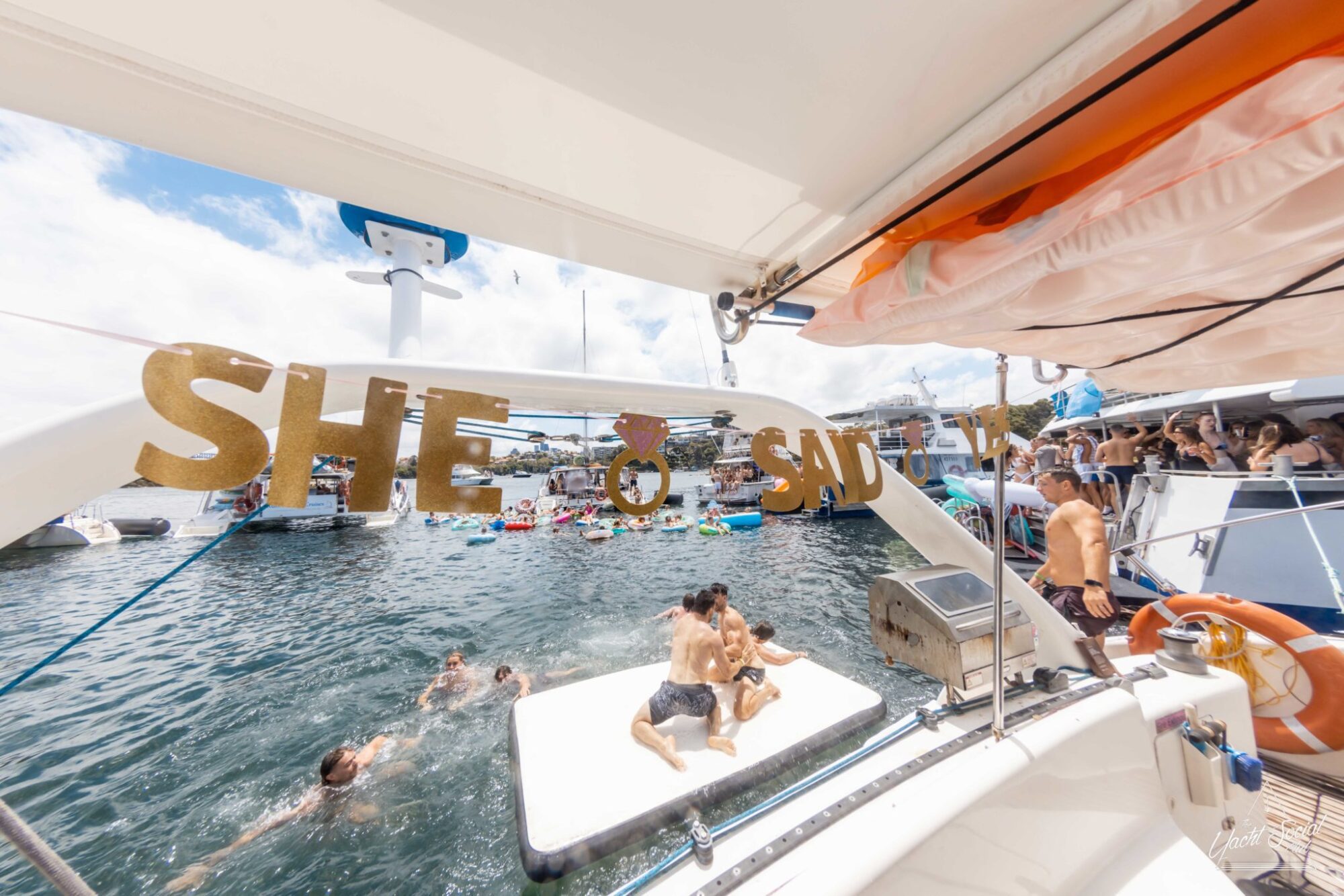 Several people are gathered on and around a boat adorned with a "SHE SAID YES" sign. Some are swimming near a float, while others enjoy the private yacht charter on Sydney Harbour with life preservers in view.