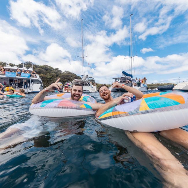 Two people floating on inflatable tubes in the water, surrounded by anchored boats and other people enjoying a sunny day at a Sydney boat party hire.