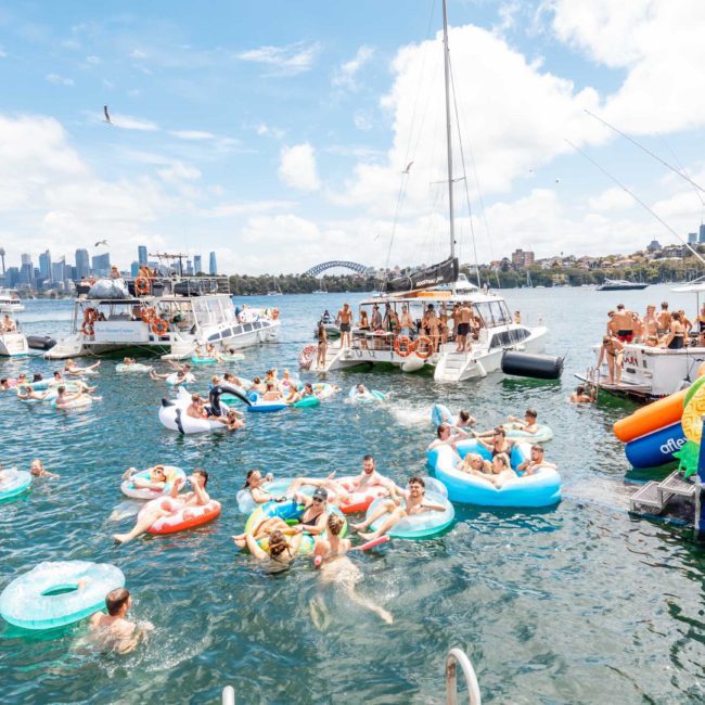 People enjoying a sunny day on the water in inflatable tubes, surrounded by boats, capturing the vibrant atmosphere of a Sydney boat party hire. A cityscape and bridge are visible in the background.