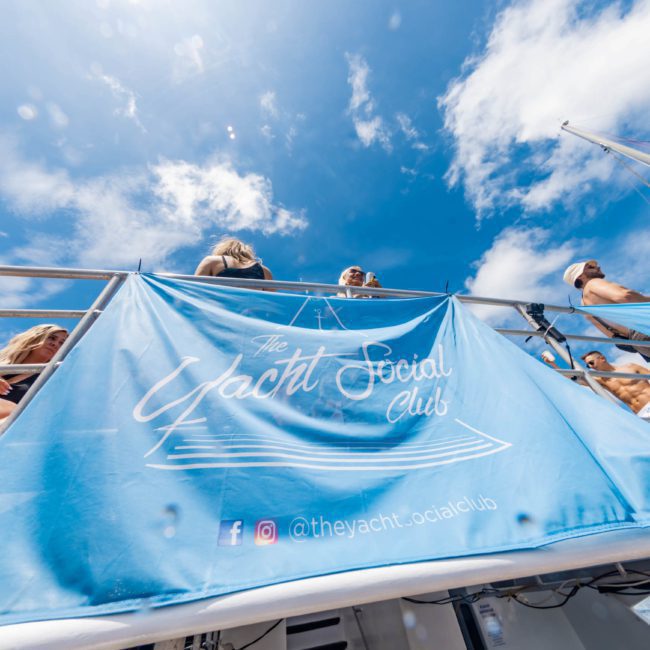People are enjoying themselves on a yacht deck adorned with a large blue banner reading "The Yacht Social Club," with a clear blue sky overhead. The DJ boat hire Sydney scene makes the event even more vibrant and unforgettable.