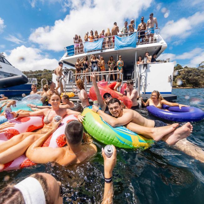 A group of people are enjoying a sunny day floating on inflatable rafts in the water next to a two-story boat, with more people standing and sitting on the boat's decks. It's the perfect scene for a Sydney boat party hire, where everyone is having a fantastic time.