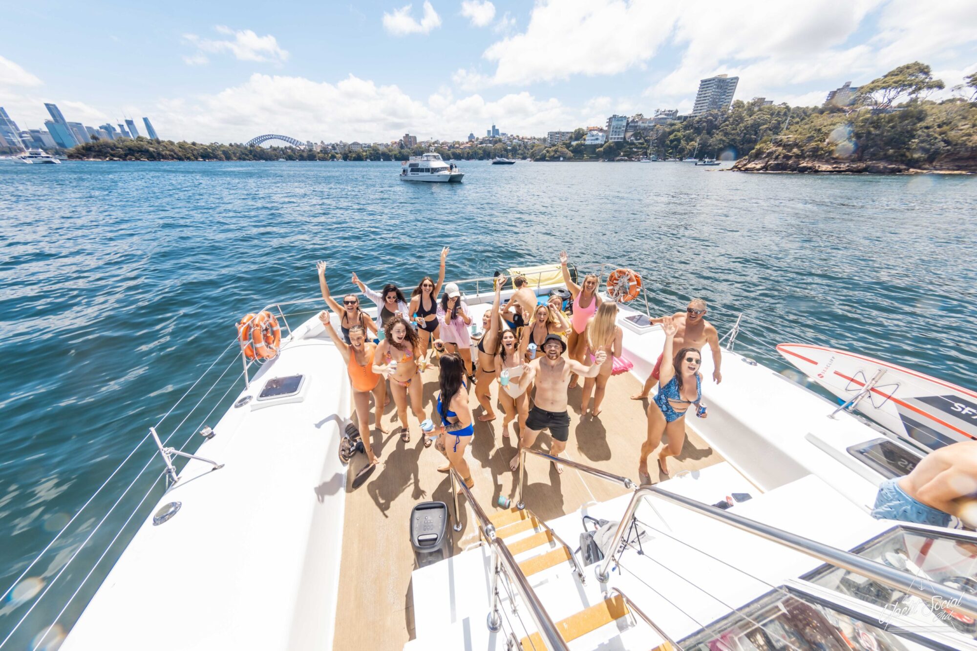 A group of people standing and posing on the deck of a luxury yacht in a sunny harbor with cityscape and several other boats in the background.