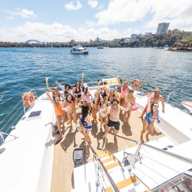 A group of people in swimwear is gathered on a yacht deck, some with their hands raised, with a view of water, trees, and city buildings in the background. Perfect for those seeking Luxury yacht hire Sydney or planning Corporate boat events Sydney.