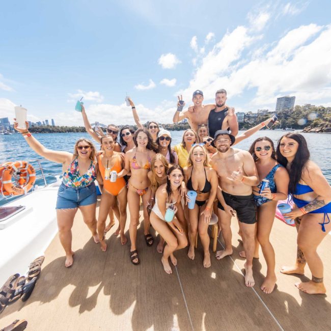 A group of people dressed in swimsuits and casual attire pose and hold drinks on a boat, with a city skyline and clear blue sky in the background, enjoying a Sydney boat party hire.
