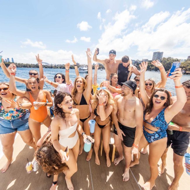 A group of people in swimsuits is gathered on a boat, smiling and raising their arms in celebration, with a city skyline and trees in the background. Perfect for corporate boat events or DJ boat hire in Sydney.