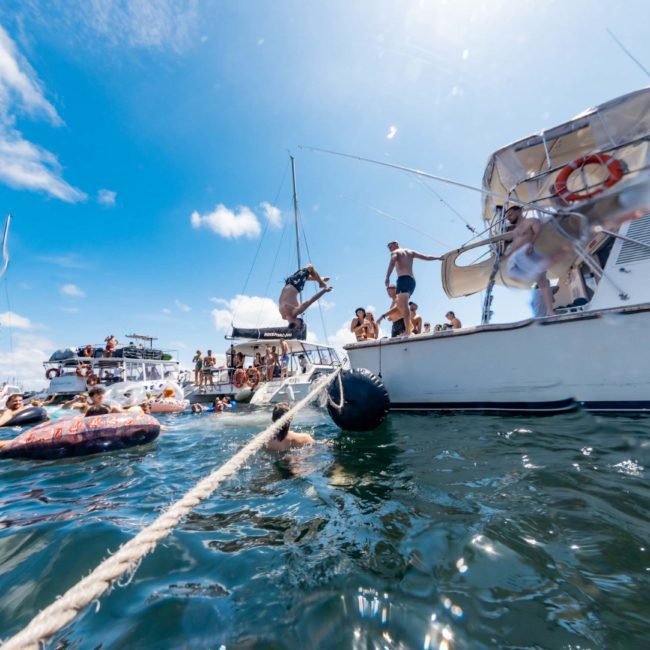People are enjoying a sunny day on boats and in the water, with some swimming and others relaxing on inflatables near the boats. It’s the perfect scene for a DJ boat hire Sydney experience, making it an unforgettable day under the sun.