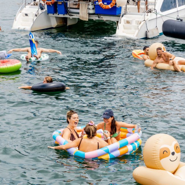 People enjoying a day on the water, some floating on inflatables including a small pool and a monkey-shaped float, near a docked boat with others on board, showcasing the fun possibilities of luxury yacht hire Sydney.