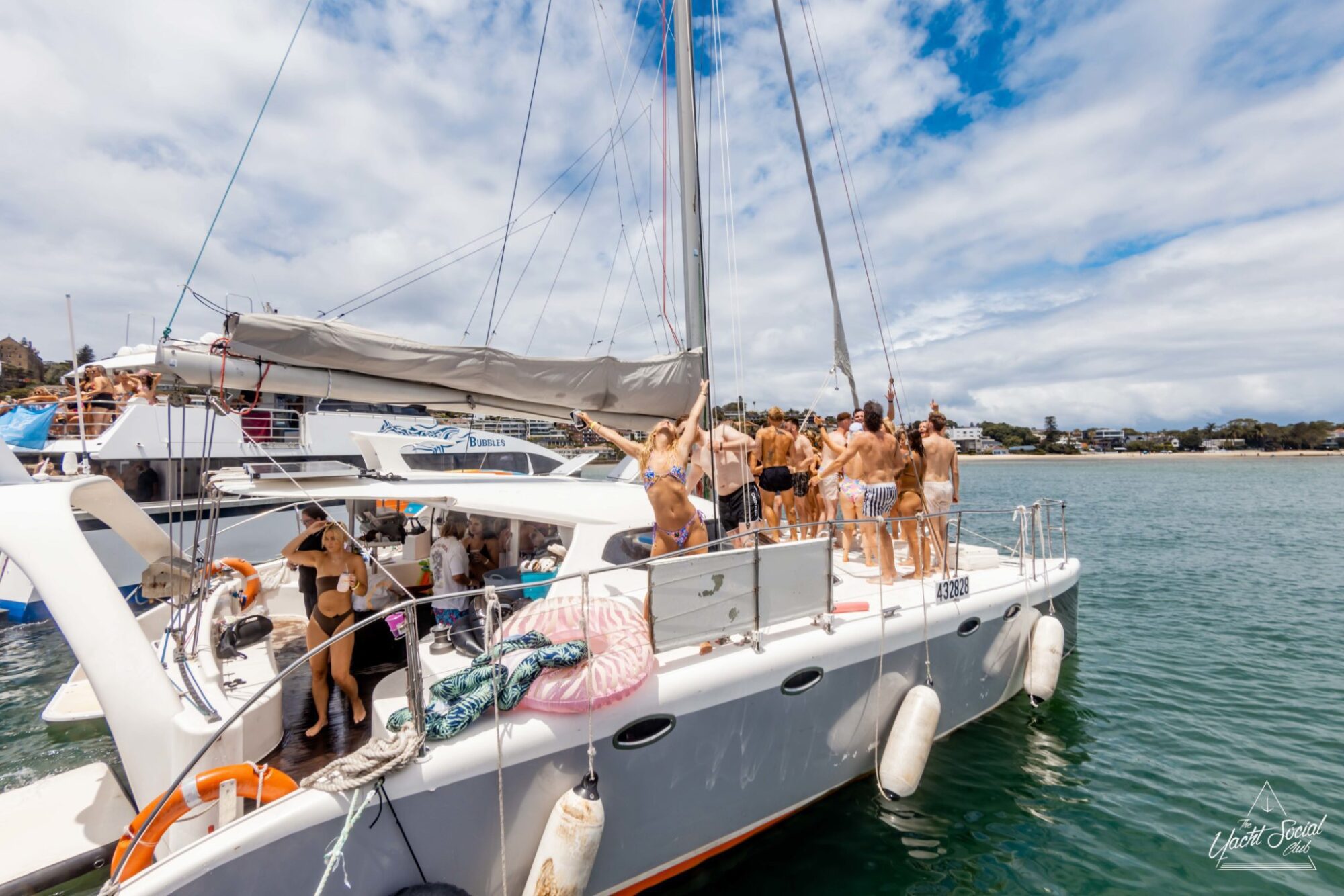 A group of people in swimwear are gathered on a sailing catamaran on the water under a partly cloudy sky, enjoying a lively catamaran party in Sydney.