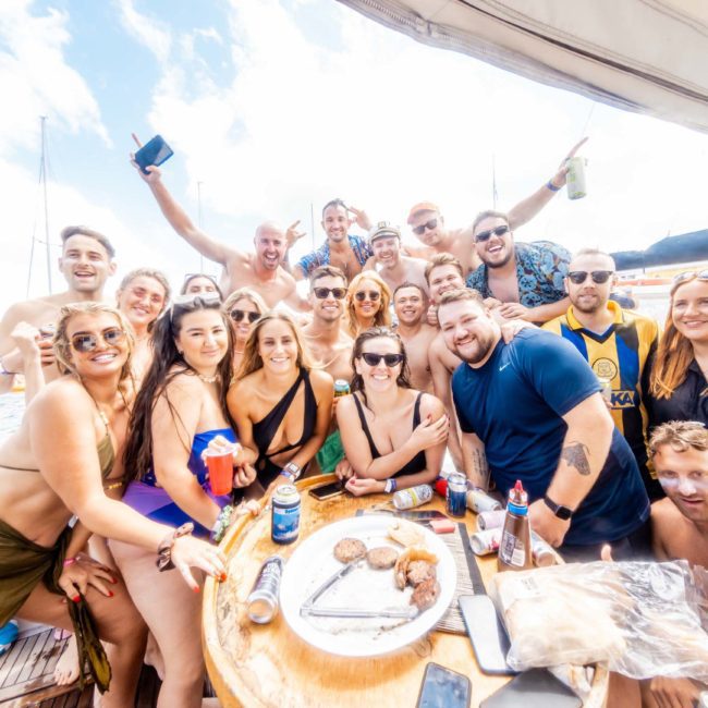 A group of people in swimwear gathered on a boat, smiling and posing with drinks. Food items are visible on a round table in the center. The background shows clear skies and water, setting the perfect scene for a Sydney boat party hire.