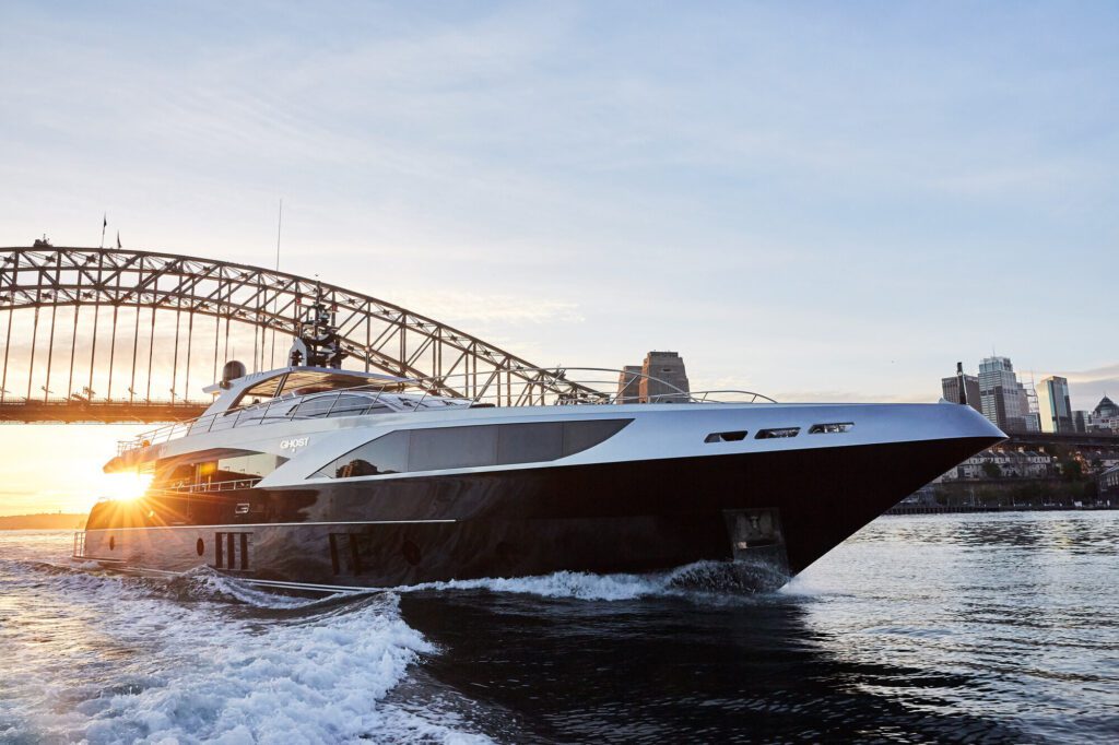 A sleek yacht cruises through the water near a large arched bridge at sunset. The sun's rays peek from behind Sydney Harbour Bridge, casting a warm glow on the scene. City buildings are visible in the background.