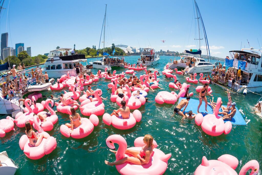 A vibrant scene of people celebrating on the water, surrounded by numerous pink flamingo pool floats. Several boats are docked nearby, and the background shows a bustling cityscape and clear blue sky, creating a lively and festive atmosphere. It's the epitome of Sydney Boat Parties.