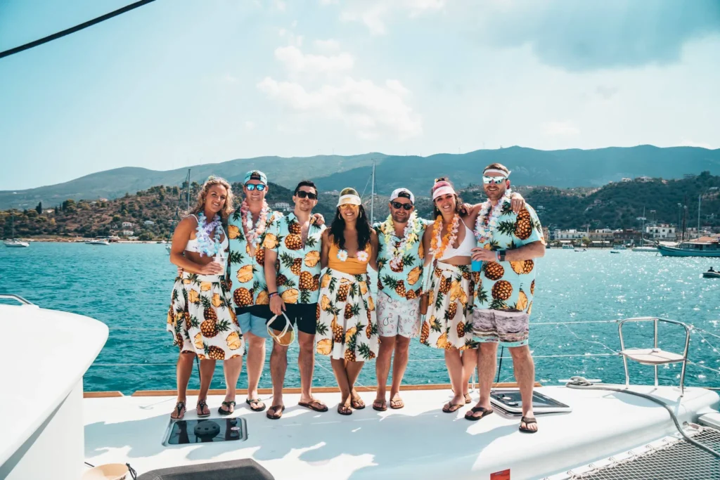 A group of seven people stands together on a boat against a backdrop of blue water and distant hills. They are all wearing matching pineapple-themed outfits and various sun hats and sunglasses, smiling and enjoying the sunny day with The Yacht Social Club Sydney Boat Hire.