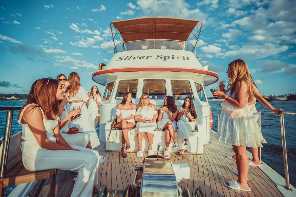 A group of women dressed in white outfits are socializing on the deck of a Sydney yacht named "Silver Spirit." They are surrounded by clear skies and calm waters, with some sitting and others standing, enjoying drinks and conversation at the corporate event.