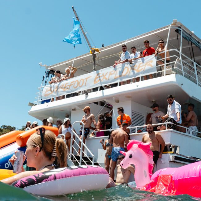 A lively scene of people enjoying a sunny day on a two-story party boat. Individuals are swimming and floating on inflatable pool toys, including a large pink unicorn. The boat's deck is crowded with people socializing amidst colorful banners and flags, creating an unforgettable summer party.