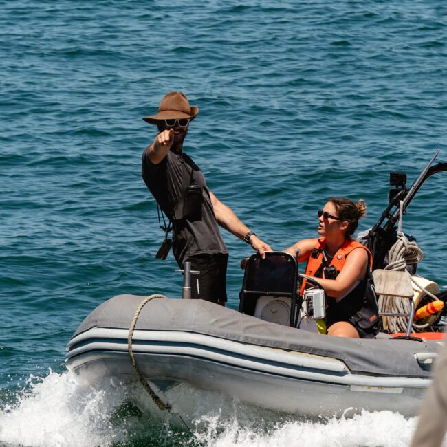 Two people are in an inflatable boat on a sunny day. One, wearing a brown hat, stands and points toward something in the distance. The other, donning sunglasses and a life vest, is seated at the controls of the boat. The peaceful water gleams under the bright sunshine.