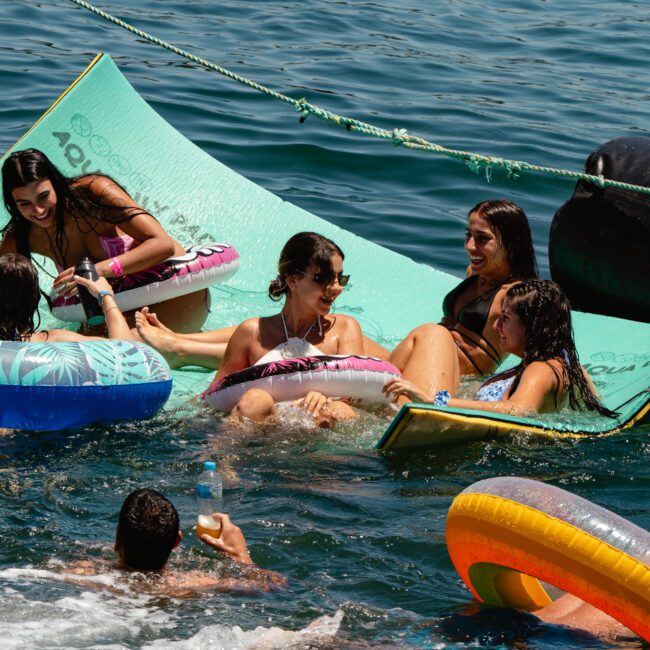 A group of people relaxing on floating mats and inflatable rings in the water next to a boat. The sunny day highlights the crystal-clear water and the joyful atmosphere. Some are talking and smiling while enjoying the refreshing swim under the sun, savoring every delightful moment.