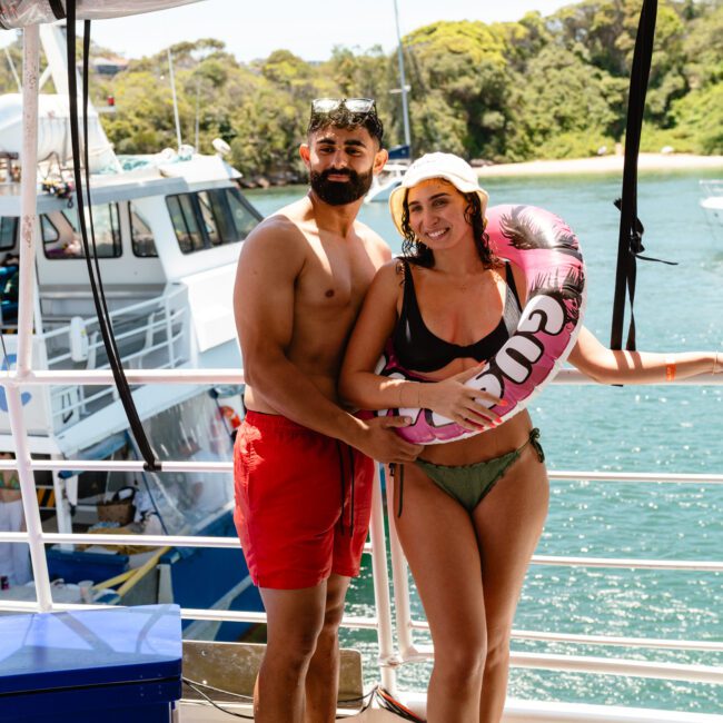 A man in red swim trunks and snorkel mask stands next to a woman in a bikini holding a pink floatation ring on a boat. The picturesque background features another boat and lush green trees by the scenic waterfront. The woman smiles while the man has a neutral expression, ready for adventure.