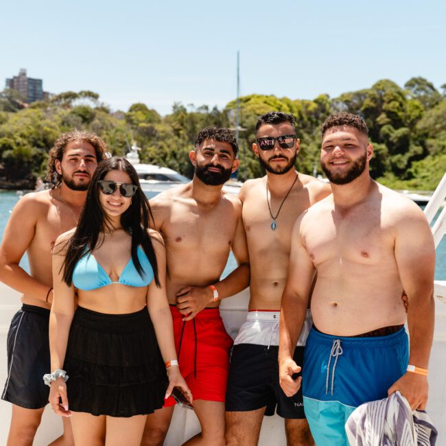 Five people, four men and one woman, posing on a boat. The woman is wearing sunglasses, a blue bikini top, and a black skirt; the men are shirtless and wearing swim trunks. They are enjoying a sunny day with greenery and sailboats visible in the background.