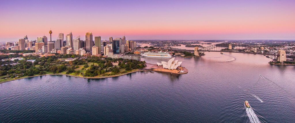 Aerial view of Sydney, Australia, at dusk. The Sydney Opera House and Sydney Harbour Bridge are prominently visible. The city skyline is bathed in pink and purple hues, with boats cruising in the harbor. Among them, a vessel from The Yacht Social Club Event Boat Charters glides through the water effortlessly.