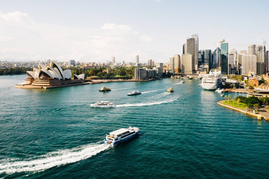 Aerial view of Sydney Harbour featuring the iconic Sydney Opera House on the left, several boats cruising the water, including luxury yacht rentals, and the city skyline with tall buildings under a clear sky in the background.