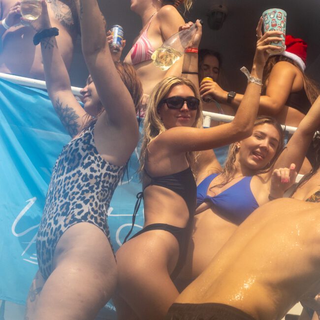 A group of people in swimsuits are partying on a boat. Four women are in the foreground, dancing and holding drinks. Other partygoers are seen in the background under a banner that reads, "Summer Bash." The scene appears joyful and energetic, capturing the essence of a fun-filled celebration.