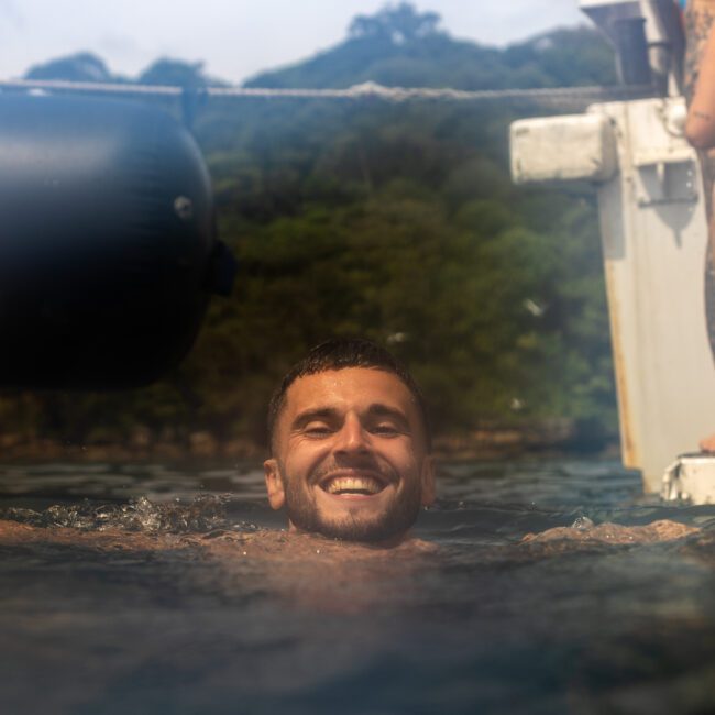 A man smiles and gives two thumbs-ups while swimming in the ocean near a boat, his short hair and beard glistening in the sun. In the background, another person in a black bikini climbs a ladder onto the boat. Lush greenery is visible in the distance, enhancing the serene setting.