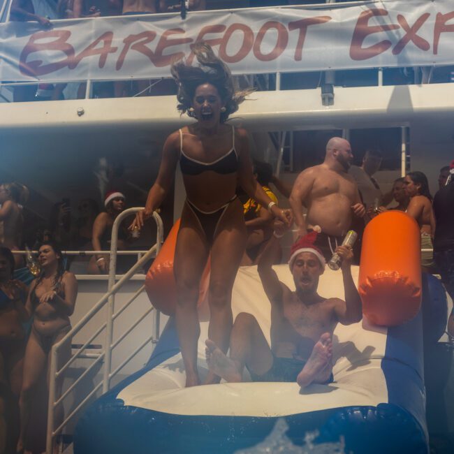 A woman in a bikini jumps off a boat while a man slides down an inflatable slide in front of her. The boat is crowded with people in swimwear, some wearing Santa hats, and a banner that reads "Barefoot Ex." The festive atmosphere is lively and celebratory.