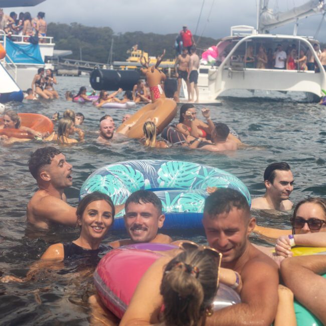 A large group of people are enjoying themselves in the crystal-clear water, surrounded by inflatable rafts and toys. Yachts and boats are anchored nearby. The scene is festive with people swimming, floating, and interacting with each other under a cloudy sky.