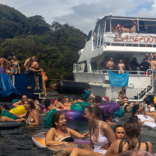 A lively scene of people enjoying a pool party on a sunny day. Many are on inflatable floats in the water, while others are on boats, including a large one labeled "Barefoot Explorer." Trees and a clear sky are visible in the background, adding to the festive atmosphere.