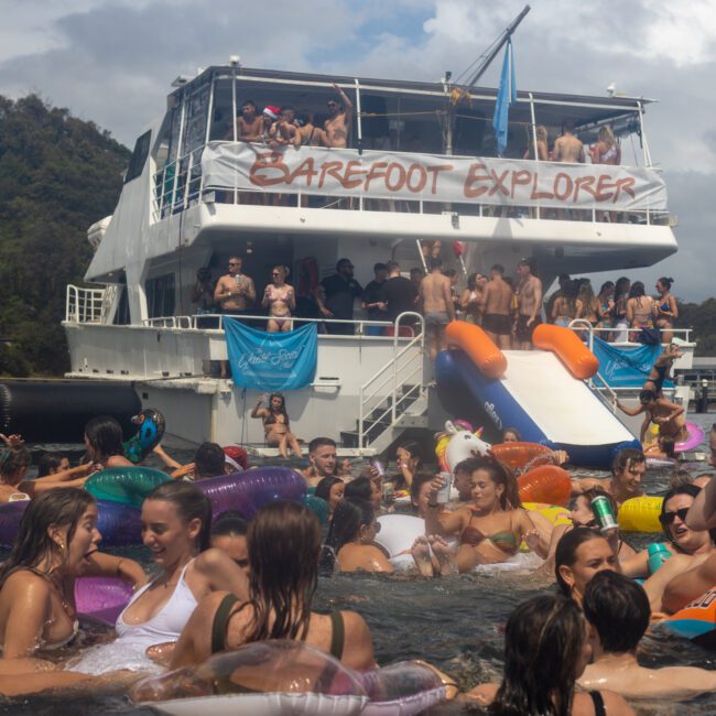 A large group of people enjoy a party on and around a boat named "Barefoot Explorer." The boat, anchored near a picturesque forested area, has a slide and many colorful inflatables in the water. People are swimming, chatting, and having fun on this vibrant sunny day.