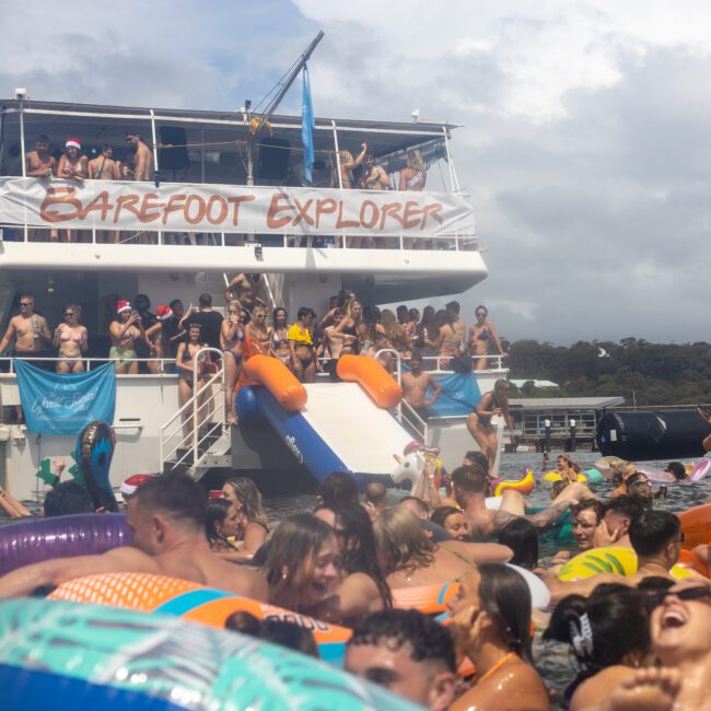 A large group of people is enjoying a sunny day on a boat and in the water. The boat, named "Barefoot Explorer," has a slide extending into the water. People are lounging on inflatable rafts and tubes, celebrating with music and having fun. The atmosphere is lively and festive.