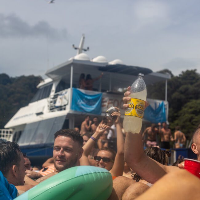 A lively group of people enjoy a sunny day in the water near a boat. One man in the foreground holds up a bottle and cheers, while others around him smile and relax on floaties. The boat in the background is crowded with partygoers under a clear blue sky. Trees add natural beauty to this festive scene.