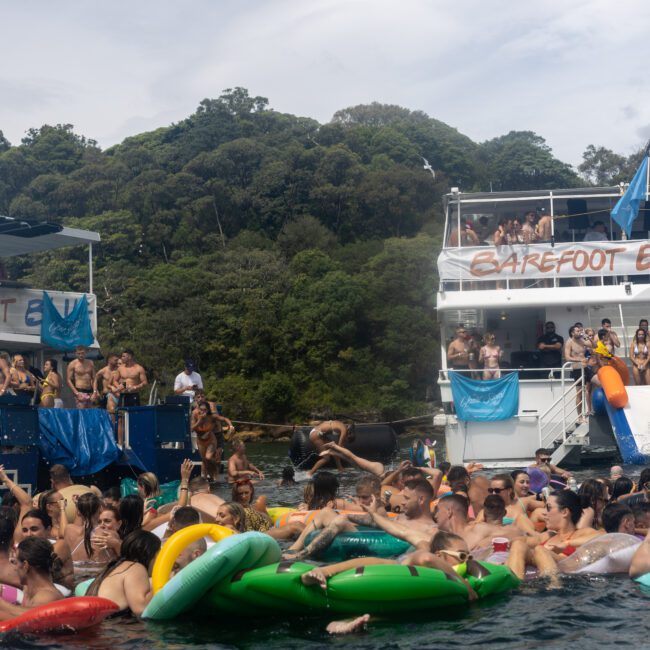 A lively scene of people enjoying a party on water floats between two boats named "Barefoot Explorer." The crowd is in colorful swimwear, with some on slides descending into the shimmering water. The boats are set against a backdrop of lush green trees and picturesque hills.