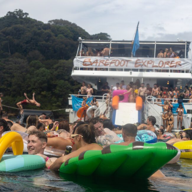 A lively scene of people enjoying a party on the water, with many floating on inflatable rafts in the foreground and a large boat labeled "Barefoot Explorer" in the background. The backdrop features lush green trees and a picturesque, partially cloudy sky.