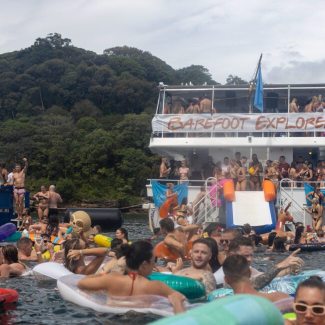 A lively party scene with numerous people swimming and floating on inflatables in the water. In the background, a double-deck boat named "Barefoot Explorer" is crowded with partying people. A fun slide from the boat leads into the water under the watchful eye of forested hills.