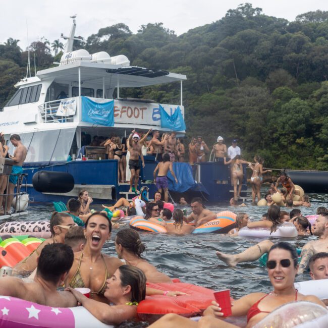 A lively boat party scene with numerous people enjoying themselves both on boats and in the water. Inflatable floats of various shapes and colors surround the boats. Many are swimming, socializing, and basking in the festive atmosphere as forested hills create a picturesque backdrop.