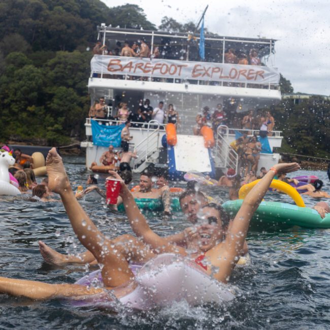 A group of people having fun in a lake festively decorated with inflatables, with a large boat labeled "Barefoot Explorer" in the background. Some splash in the water while others enjoy ice-cold drinks and lively music both onboard and around the boat.