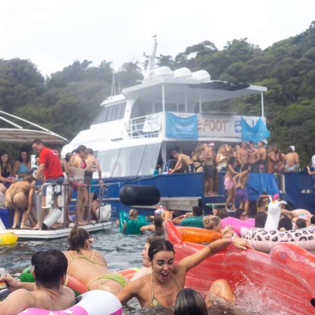 A lively group of people are enjoying a party on colorful inflatable floaties in the water near a large boat. The background shows more people on the boat, with trees and a cloudy sky visible. The mood is festive and energetic, emphasizing social interaction and fun.