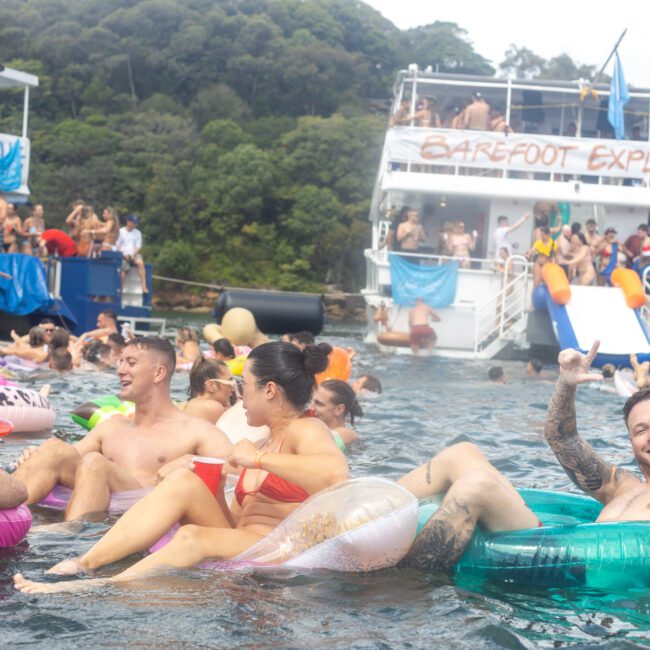 A lively crowd enjoys a floating party in a scenic bay. People relax on colorful inflatables in the water, holding drinks and laughing. In the background, a large boat labeled "Barefoot Explorer" hosts more partygoers and features an inflatable slide into the water.